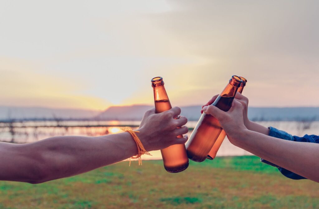 A group of Asian friends tourist drinking alcoholic beer and playing guitar together with happiness in Summer while having camping near lake at sunset
