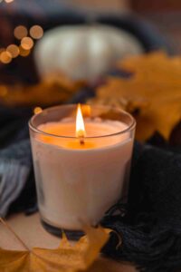 A lit candle in a glass jar with a cozy, autumnal background featuring blurred orange leaves and soft lighting